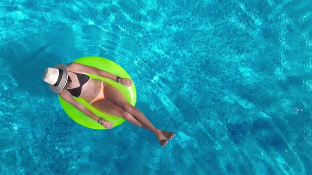 Aerial drone view of young woman in hat relaxing in swimming pool, top view of beautiful girl in bikini floats on inflatable ring donut and has fun in water, tropical vacation on holiday resort