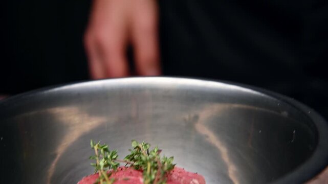 Chef prepares raw beef steak with fresh thyme in stainless steel bowl