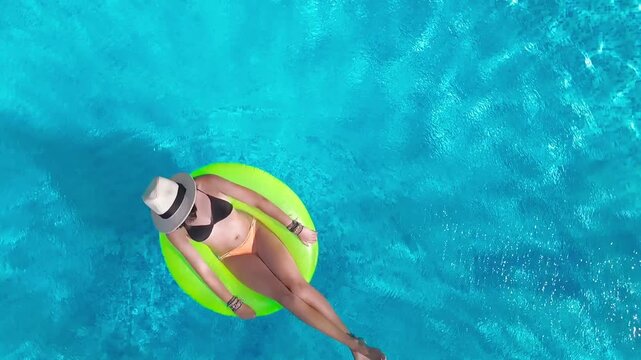 Aerial drone view of young woman in hat relaxing in swimming pool, top view of beautiful girl in bikini floats on inflatable ring donut and has fun in water, tropical vacation on holiday resort