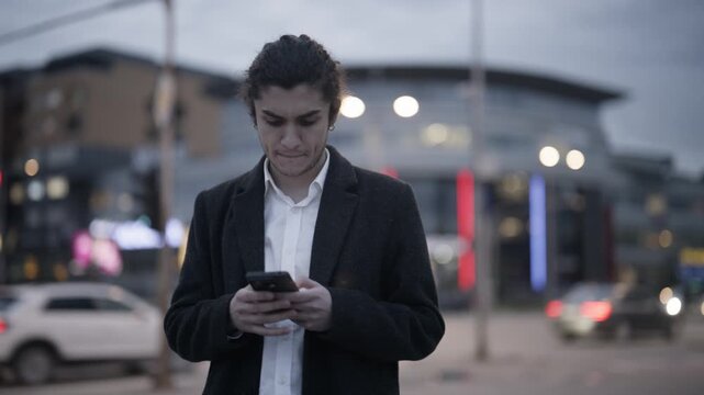 A young man in a business coat stands on a city street, initially looking confused before finding content on his smartphone and smiling.