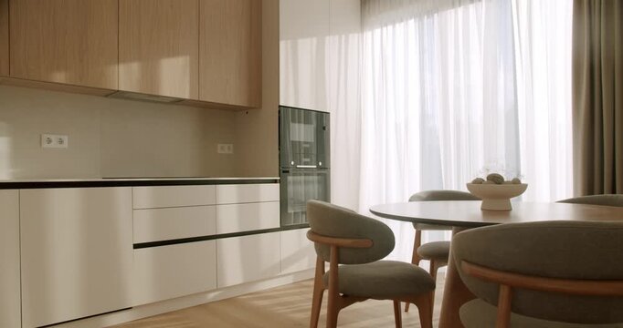 Bright and spacious kitchen interior with minimalist wooden cabinets and a contemporary dining set next to a large window with sheer curtains, bathed in beautiful morning sunlight