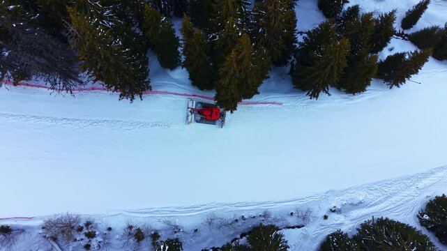 A snow groomer moves along a ski trail in a forested mountain area. The machine pushes snow to create a smooth surface for skiers. Tall trees surround the scene, adding to the winter landscape.