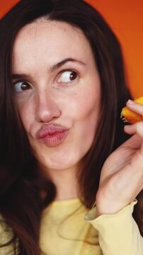 Vertical close up of brunette woman biting lemon slice and pouting lips on orange background. Beautiful girl tasting sour citrus fruit and looking at camera. Healthy lifestyle and fun concept.