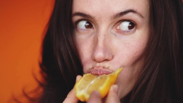 Horizontal extreme close up of woman biting a sour lemon slice and making a funny face on orange background. Brunette girl tasting citrus fruit and squinting eyes. Healthy lifestyle concept