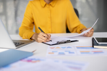 Businesswoman signing a document on a clipboard, working at a desk with a laptop, tablet, and...