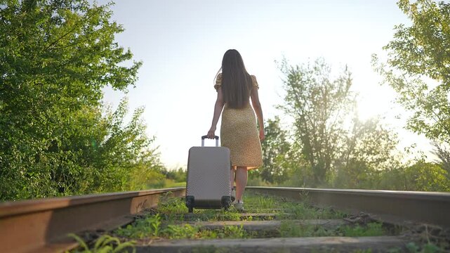 Young woman walks along railroad tracks pulling suitcase. Summer sun sets. Female traveler drags luggage on railway. Girl walking with suitcase starts journey. Adventure calls. Sun shines bright.