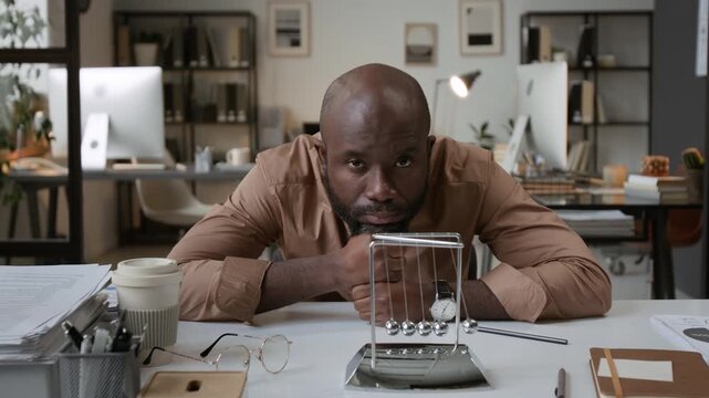 Zoom in portrait shot of pensive bearded African American man resting his head on fists and looking at camera while sitting at office desk with Newtons cradle