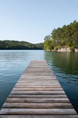 Fototapeta premium Wooden dock leading into calm lake with surrounding pine forest and rocky shoreline beneath a clear sky