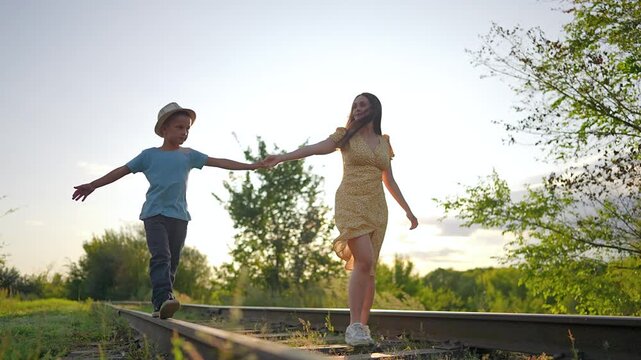 Happy mother and son walk on railroad tracks at sunset. Mother holding hands with son helps him balance on rail. Family enjoys summer walk on railroad. Sun shines bright on happy mother and son.