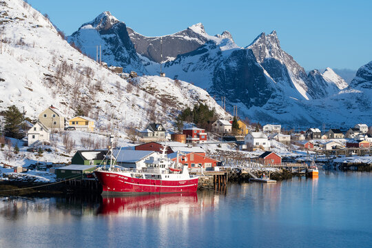 Lofoten, Norway - 28.03.2024: Fishing boat moored at harbor with snowy mountains and coastal village in background