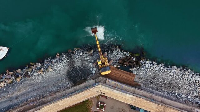 A machine lifts and moves gravel and rocks along the shore next to a body of water. Boats are visible nearby, and rocks line the water's edge under a clear sky.