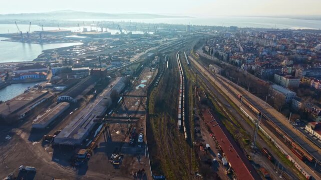 An industrial area with rail tracks, buildings, and several cranes along the waterfront. The setting includes a glimpse of the port and nearby structures.