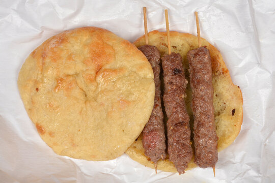 Three traditional grilled sis cevapi meat skewers laid out on an open fresh lepinja flatbread placed on white paper