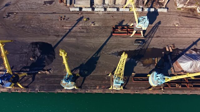 Cranes lift cargo from containers at a shipping dock next to the water. Workers are present on the dock managing the operation in the afternoon.