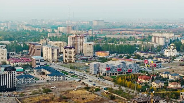 Drone trucking right over urban mix: modern low-rise buildings with colorful accents, unfinished high-rises, cranes, panel apartments, greenery, hazy smog, distant TEC chimneys at dusk.