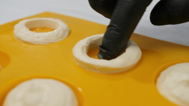 Confectioner hand in gloves puts mango filling into cream in forms on tray closeup. Master chef cooks delicious mochi cakes with exotic fruits at countertop. Japanese dessert preparation