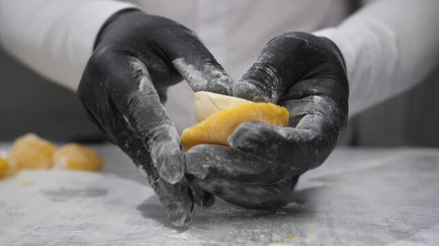 Confectioner in black gloves shapes tasty mango mochi with yellow rice dough in bakery closeup. Japanese chef hands cook delicious cakes with tropical fruits over countertop