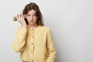 Young woman in yellow sweater holds smartphone to ear with confused expression, isolated on white...