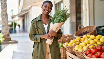 Obraz premium Smiling Black woman holding a bouquet of fresh herbs at an outdoor market. Happy female shopper standing near fruit crates with organic produce. Healthy lifestyle and sustainable shopping concept