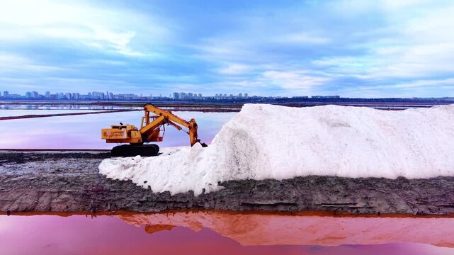 Side view of a yellow hydraulic excavator on a salt ridge with city buildings in the background and pink water reflections. Industrial excavator processing sea salt at evaporation site.
