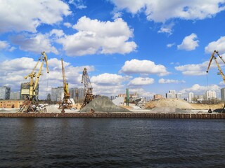 Fototapeta premium Industrial river cargo port with cranes, sand piles and city skyline under blue sky with clouds.