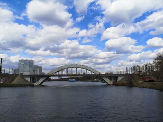 Fototapeta premium Urban arched bridge spanning a calm river with modern buildings nearby and dramatic clouds in a blue sky cityscape.