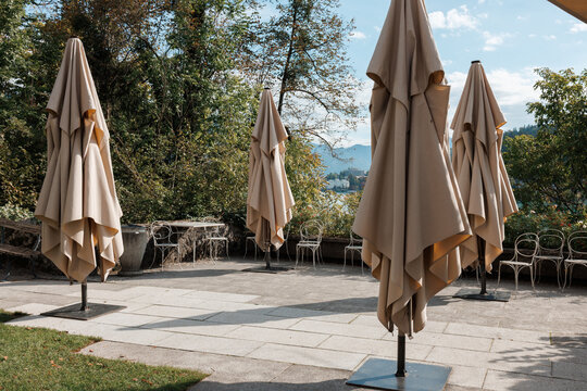Large beige sun umbrellas remain closed on an empty stone cafe terrace overlooking the trees and water of Lake. Stock photo