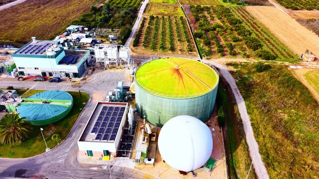 Green biogas storage tank, white spherical gas holder, and solar photovoltaic array at a renewable energy facility. Close up aerial view of biogas digester and solar panels.