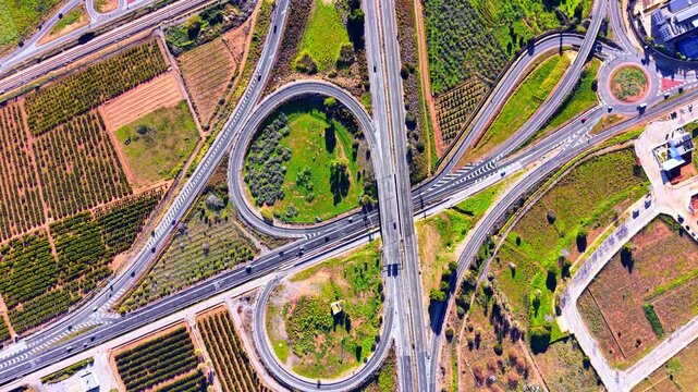 Aerial shot of a multi-level road junction with loop ramps, green spaces, and surrounding orange orchards. Top down view of a cloverleaf highway interchange.