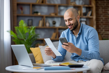 Young man smiling in a cozy home office, holding a bill and smartphone while managing online...