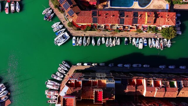 Top down aerial shot of white yachts and boats docked along a pier with red tiled roofs. Boats moored in marina with turquoise water.