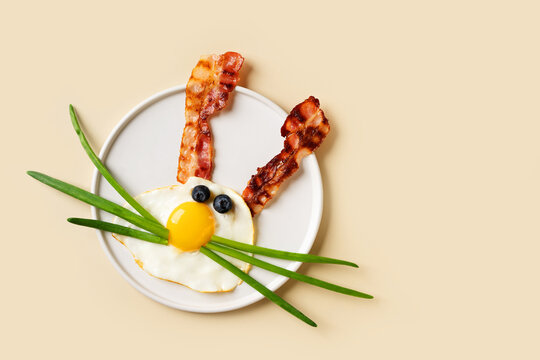 Easter concept with funny fried egg bunny and becon on plate on beige background, top view