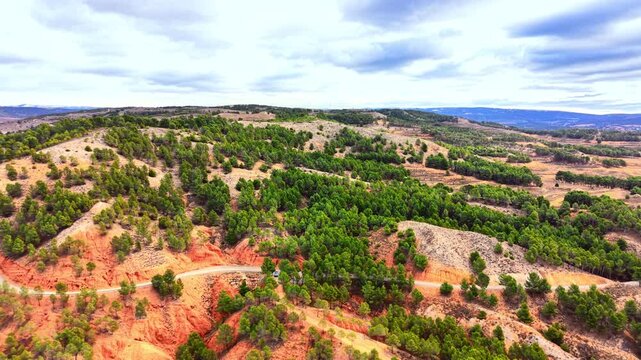 Aerial view of a curved asphalt road passing through red soil hills covered with green pine trees under a overcast sky. Winding road through red clay hills.