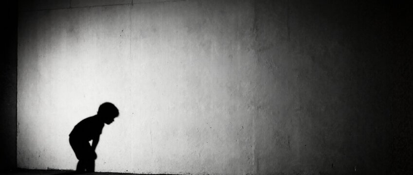 Silhouette of sad lonely child crouching against concrete wall. Black and white concept of childhood depression and isolation. Copy space for text