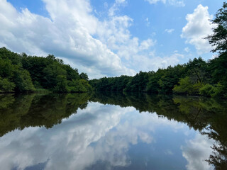 Obraz premium Serene landscape featuring calm river reflecting blue sky and clouds, surrounded by lush green trees and foliage in a tranquil natural setting