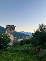 Ancient stone watchtowers surrounded by lush greenery and residential buildings with mountains in the background during twilight in a rural setting