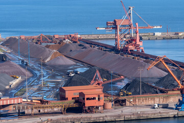 A wide view of the mineral storage area at the Port of Gijon, Asturias, featuring large piles of coal and iron ore with industrial conveyors and port cranes.
