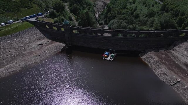 An aerial view of a dam with a low water level. A small platform with containers floats on the dark water.
