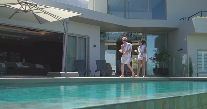 Diverse couple in swimwear exiting sliding glass doors, unfolding striped towel by pool, settling