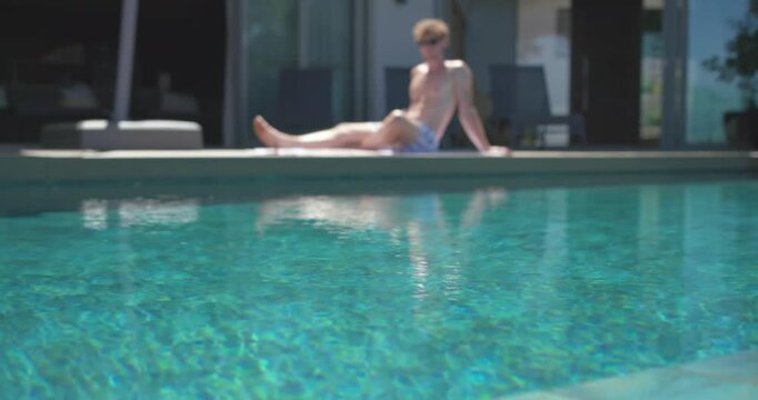 Camera pulling back shirtless man shifting lowering torso and adjusting sunglasses relaxing by pool
