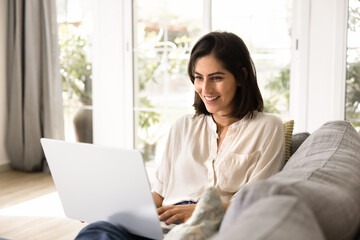 Happy young Latin woman resting on soft couch, typing on laptop, browsing social media, using Internet for communication, shopping. Freelancer working on project from home