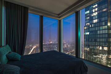 A bedroom located in a tall building offers a view of the city skyline at night. The lights from the streets and buildings create a vibrant atmosphere outside the large windows © Alhim