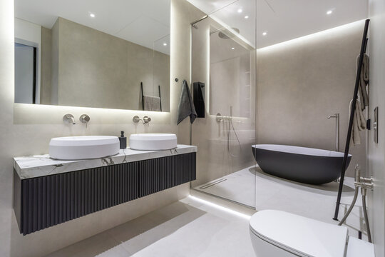 A bathroom features two white sinks on a wall-mounted vanity. A freestanding tub sits against the wall. Soft lights create a bright setting. Towels hang nearby, and sleek fixtures are visible