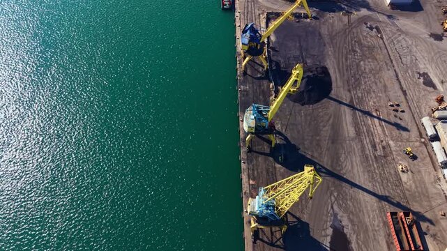Cranes are positioned on a dock looking over a blue sea. Workers are moving materials around the area in a busy port setting.