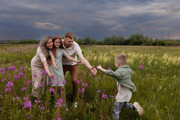 Caucasian Family Playing Together In Meadow, Parents Encouraging Young Child To Run Across...