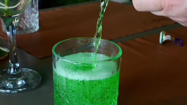 a person pouring a bright, neon green carbonated beverage&mdash;classic Georgian Tarhun (tarragon soda)&mdash;from a green bottle into a clear glass, creating a fizzy, bubbly foam on a restaurant table.