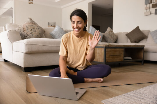Cheerful Latina woman sitting cross-legged on yoga mat in front of laptop, smiling and waving at screen during online yoga or meditation practice from home. Modern tech usage, virtual workouts session