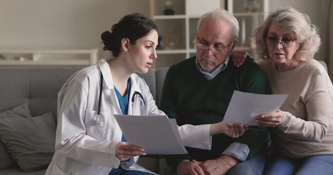 Young adult female doctor explaining medical test results or diagnosis to senior 70s couple, handing over prescription, discussing together insurance terms during home visit consultation. Health care