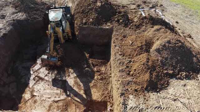 Aerial drone footage of a backhoe loader excavating a foundation trench and moving soil around an active construction site.