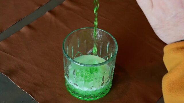 a person pouring a bright, neon green carbonated beverage&mdash;classic Georgian Tarhun (tarragon soda)&mdash;from a green bottle into a clear glass, creating a fizzy, bubbly foam on a restaurant table.